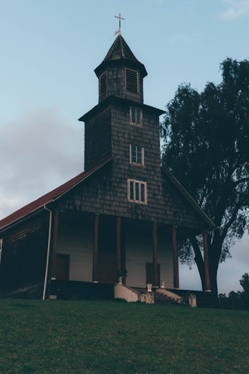 A rustic wooden church with a shingled roof stands on a grassy hill. It has a tall bell tower topped with a cross, and a large tree is visible to the side. The sky is cloudy, adding a serene and peaceful atmosphere.