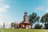 A quaint wooden church stands on a grassy hill, with trees surrounding it under a clear blue sky. The structure features a steeple with a cross on top and red accents along the roofline and doorway.