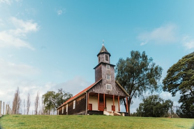A quaint wooden church stands on a grassy hill, with trees surrounding it under a clear blue sky. The structure features a steeple with a cross on top and red accents along the roofline and doorway.