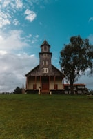 A rustic, multi-story wooden church stands on a grassy hill, surrounded by a few trees. The building features a tall steeple with a cross on top and a symmetrical design with wooden doors and windows. The sky is partly cloudy, creating a tranquil and serene atmosphere.