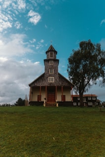 A rustic, multi-story wooden church stands on a grassy hill, surrounded by a few trees. The building features a tall steeple with a cross on top and a symmetrical design with wooden doors and windows. The sky is partly cloudy, creating a tranquil and serene atmosphere.