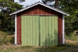 A small wooden shed with a red exterior and a large green door stands amidst a natural setting. The structure is surrounded by grass and trees, with sunlight casting dappled shadows on the scene.