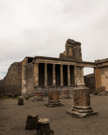 Ancient ruins featuring large stone columns and remnants of a historical building. The structures are set in a dusty, open area surrounded by aged walls, with intricate detailing evident on the columns.