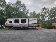 A travel trailer labeled 'Autumn Ridge' is parked on a gravel lot. The trailer is positioned near a wooden deck and surrounded by lush green trees under a cloudy sky. A tractor is partially visible behind the trailer.