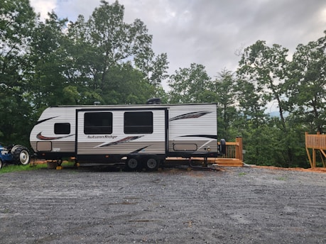 A travel trailer labeled 'Autumn Ridge' is parked on a gravel lot. The trailer is positioned near a wooden deck and surrounded by lush green trees under a cloudy sky. A tractor is partially visible behind the trailer.