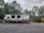 A travel trailer labeled 'Autumn Ridge' is parked on a gravel lot. The trailer is positioned near a wooden deck and surrounded by lush green trees under a cloudy sky. A tractor is partially visible behind the trailer.