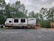 A travel trailer labeled 'Autumn Ridge' is parked on a gravel lot. The trailer is positioned near a wooden deck and surrounded by lush green trees under a cloudy sky. A tractor is partially visible behind the trailer.