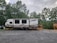 A travel trailer labeled 'Autumn Ridge' is parked on a gravel lot. The trailer is positioned near a wooden deck and surrounded by lush green trees under a cloudy sky. A tractor is partially visible behind the trailer.