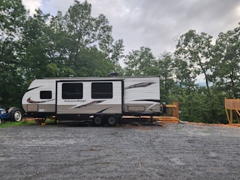 A travel trailer labeled 'Autumn Ridge' is parked on a gravel lot. The trailer is positioned near a wooden deck and surrounded by lush green trees under a cloudy sky. A tractor is partially visible behind the trailer.