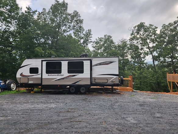 A travel trailer labeled 'Autumn Ridge' is parked on a gravel lot. The trailer is positioned near a wooden deck and surrounded by lush green trees under a cloudy sky. A tractor is partially visible behind the trailer.