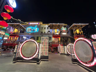 A vibrant urban scene features a historic architectural facade adorned with colorful neon lights, including a bright blue vehicle graphic and decorative lanterns. The structure is surrounded by large circular installations framed with glowing red lights, creating a lively yet nostalgic ambiance.