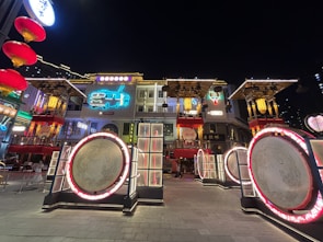 A vibrant urban scene features a historic architectural facade adorned with colorful neon lights, including a bright blue vehicle graphic and decorative lanterns. The structure is surrounded by large circular installations framed with glowing red lights, creating a lively yet nostalgic ambiance.