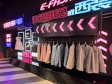 A stylish clothing display with suits and dresses hung against a backdrop of vibrant neon lights and decorative signage. Several suits are lined on a rack, with a white dress and shoes on display. The background features a modern, neon aesthetic with text in multiple languages.