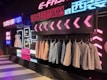 A stylish clothing display with suits and dresses hung against a backdrop of vibrant neon lights and decorative signage. Several suits are lined on a rack, with a white dress and shoes on display. The background features a modern, neon aesthetic with text in multiple languages.
