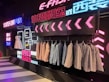 A stylish clothing display with suits and dresses hung against a backdrop of vibrant neon lights and decorative signage. Several suits are lined on a rack, with a white dress and shoes on display. The background features a modern, neon aesthetic with text in multiple languages.