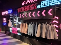 A stylish clothing display with suits and dresses hung against a backdrop of vibrant neon lights and decorative signage. Several suits are lined on a rack, with a white dress and shoes on display. The background features a modern, neon aesthetic with text in multiple languages.