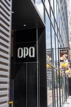 A modern building facade featuring large reflective glass panels and a black section with white letters. Visible reflections on the glass add depth and urban context. Nearby are signs displaying images of food and Japanese text, likely indicating a nearby cafe or restaurant.