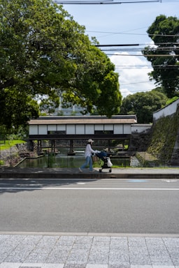 A person pushes a stroller along a sidewalk next to a wide road. In the background, a traditional-style wooden bridge spans over a body of water. Large lush trees surround the scene, casting shadows on the path. Stone walls and greenery add a tranquil, natural feel.