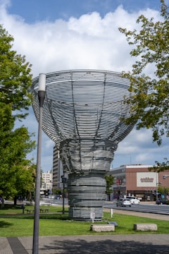 A large, metallic, spiral structure resembling a vortex is situated in a park setting with green grass and trees surrounding it. The sky is partly cloudy, and several buildings are visible in the background. Cars can be seen on a nearby road, and there is some urban street furniture such as benches present.