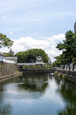 A tranquil scene featuring a waterway with reflective water, bordered by stone walls and lush green trees. A traditional Japanese architecture building with white walls and a tiered roof stands prominently. The sky is bright with scattered clouds, and the surroundings are serene and peaceful.