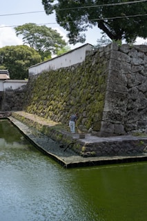 A person is cleaning a stone pathway beside an ancient stone wall with moss growing on it. The wall is adjacent to a body of water, possibly a moat, and surrounded by lush green trees and vegetation. The scene is calm and appears to be a historical or traditional setting.