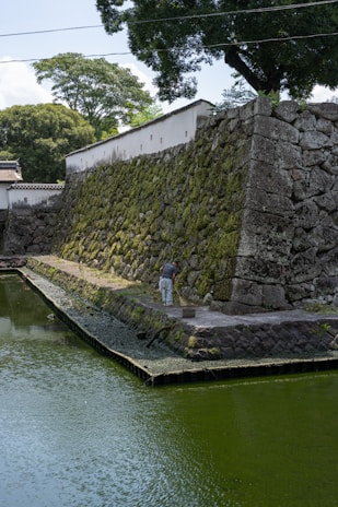A person is cleaning a stone pathway beside an ancient stone wall with moss growing on it. The wall is adjacent to a body of water, possibly a moat, and surrounded by lush green trees and vegetation. The scene is calm and appears to be a historical or traditional setting.