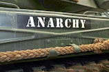 A close-up of a green metallic surface featuring the word 'ANARCHY' in bold white stencil letters. Below the text, there is a thick, twisted rope attached to the surface with metal brackets.