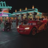 A bright red animated car character with the number 95 and eyes on the windshield drives down a nighttime street. It is surrounded by a crowd of people, including families and children, some with strollers. The background features a neon-lit diner with a retro aesthetic, showcasing bright teal and pink lights.