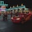 A bright red animated car character with the number 95 and eyes on the windshield drives down a nighttime street. It is surrounded by a crowd of people, including families and children, some with strollers. The background features a neon-lit diner with a retro aesthetic, showcasing bright teal and pink lights.