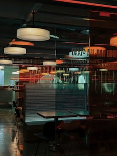Interior of a dining area in a restaurant with multiple hanging ceiling lights illuminating the space. There are several tables and chairs arranged, some of them separated by glass partitions. The environment has a mix of industrial and modern design elements with reflections on the glass surfaces, creating a layered and somewhat complex visual effect. The words 'ALL-DAY DINING' and 'DOWN TO EARTH' are partially visible on one of the glass partitions.