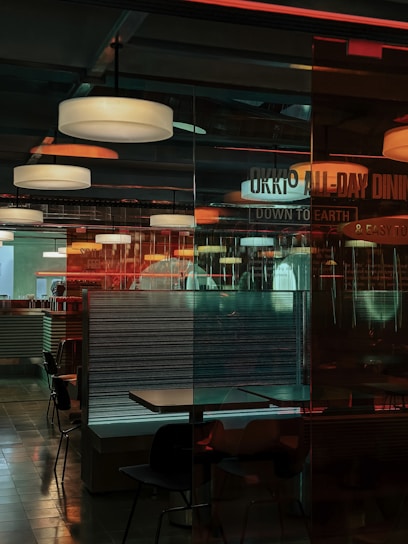 Interior of a dining area in a restaurant with multiple hanging ceiling lights illuminating the space. There are several tables and chairs arranged, some of them separated by glass partitions. The environment has a mix of industrial and modern design elements with reflections on the glass surfaces, creating a layered and somewhat complex visual effect. The words 'ALL-DAY DINING' and 'DOWN TO EARTH' are partially visible on one of the glass partitions.
