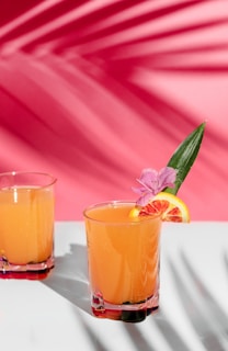 A vibrant orange drink served in a glass, garnished with a slice of orange, a pink flower, and a green leaf. Another similar glass is partially visible in the background. The setting features a bright pink backdrop with shadows of leaves creating a tropical ambiance.