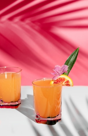 A vibrant orange drink served in a glass, garnished with a slice of orange, a pink flower, and a green leaf. Another similar glass is partially visible in the background. The setting features a bright pink backdrop with shadows of leaves creating a tropical ambiance.