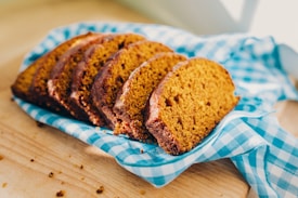 Several slices of freshly baked pumpkin bread are arranged on a light blue checkered cloth. The bread has a golden-brown crust with a soft, moist interior. Crumbs are visible on the wooden surface beneath.