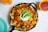 A black skillet filled with nachos topped with melted cheese, diced tomatoes, sliced avocados, sour cream, and garnished with chopped cilantro. A side dish of salsa in a yellow bowl and an orange drink are visible on the table.