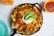 A black skillet filled with nachos topped with melted cheese, diced tomatoes, sliced avocados, sour cream, and garnished with chopped cilantro. A side dish of salsa in a yellow bowl and an orange drink are visible on the table.