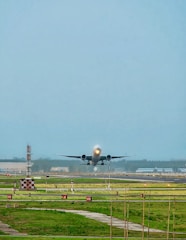 A large airplane is taking off from a runway, surrounded by green grass and airport infrastructure. The sky is clear with a light blue color, and there are some airport signs and lighting along the runway.