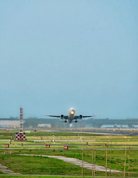A large airplane is taking off from a runway, surrounded by green grass and airport infrastructure. The sky is clear with a light blue color, and there are some airport signs and lighting along the runway.