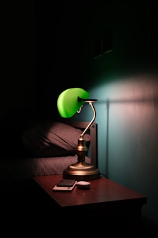 A brass desk lamp with a green shade casts a soft light on a wooden bedside table. The table holds a smartphone and a small, white device. The surrounding environment is dimly lit, with a bed featuring gray bedding partially visible.