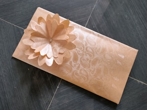 A rectangular envelope with an intricate floral pattern embossed on its surface, featuring a three-dimensional flower decoration made from the same material. The envelope is placed on a dark tiled floor.