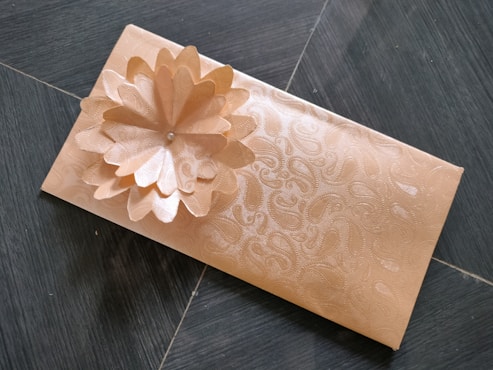 A rectangular envelope with an intricate floral pattern embossed on its surface, featuring a three-dimensional flower decoration made from the same material. The envelope is placed on a dark tiled floor.