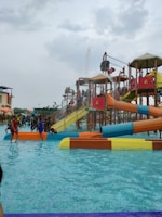 A water park scene with children and families enjoying various water slides and play structures. The colorful slides are elevated and connected by staircases and bridges. People are scattered around, some climbing the structures, while others are in the shallow pool area.