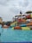 A water park scene with children and families enjoying various water slides and play structures. The colorful slides are elevated and connected by staircases and bridges. People are scattered around, some climbing the structures, while others are in the shallow pool area.