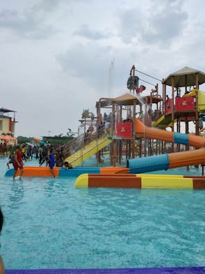 A water park scene with children and families enjoying various water slides and play structures. The colorful slides are elevated and connected by staircases and bridges. People are scattered around, some climbing the structures, while others are in the shallow pool area.