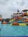 A water park scene with children and families enjoying various water slides and play structures. The colorful slides are elevated and connected by staircases and bridges. People are scattered around, some climbing the structures, while others are in the shallow pool area.