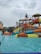 A water park scene with children and families enjoying various water slides and play structures. The colorful slides are elevated and connected by staircases and bridges. People are scattered around, some climbing the structures, while others are in the shallow pool area.
