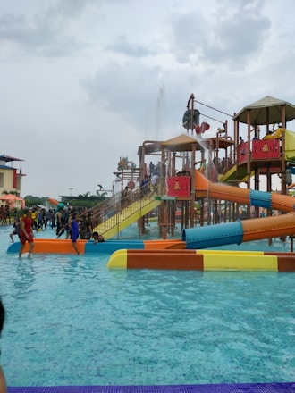 A water park scene with children and families enjoying various water slides and play structures. The colorful slides are elevated and connected by staircases and bridges. People are scattered around, some climbing the structures, while others are in the shallow pool area.