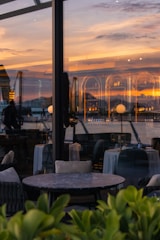 The photograph captures a serene and elegant setting in a modern glass-walled restaurant or café with empty tables. A stunning sunset reflects on the glass, casting warm hues across the scene. The view includes a body of water and distant silhouettes of buildings or hills, adding a picturesque backdrop.