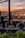The photograph captures a serene and elegant setting in a modern glass-walled restaurant or café with empty tables. A stunning sunset reflects on the glass, casting warm hues across the scene. The view includes a body of water and distant silhouettes of buildings or hills, adding a picturesque backdrop.