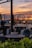 The photograph captures a serene and elegant setting in a modern glass-walled restaurant or café with empty tables. A stunning sunset reflects on the glass, casting warm hues across the scene. The view includes a body of water and distant silhouettes of buildings or hills, adding a picturesque backdrop.